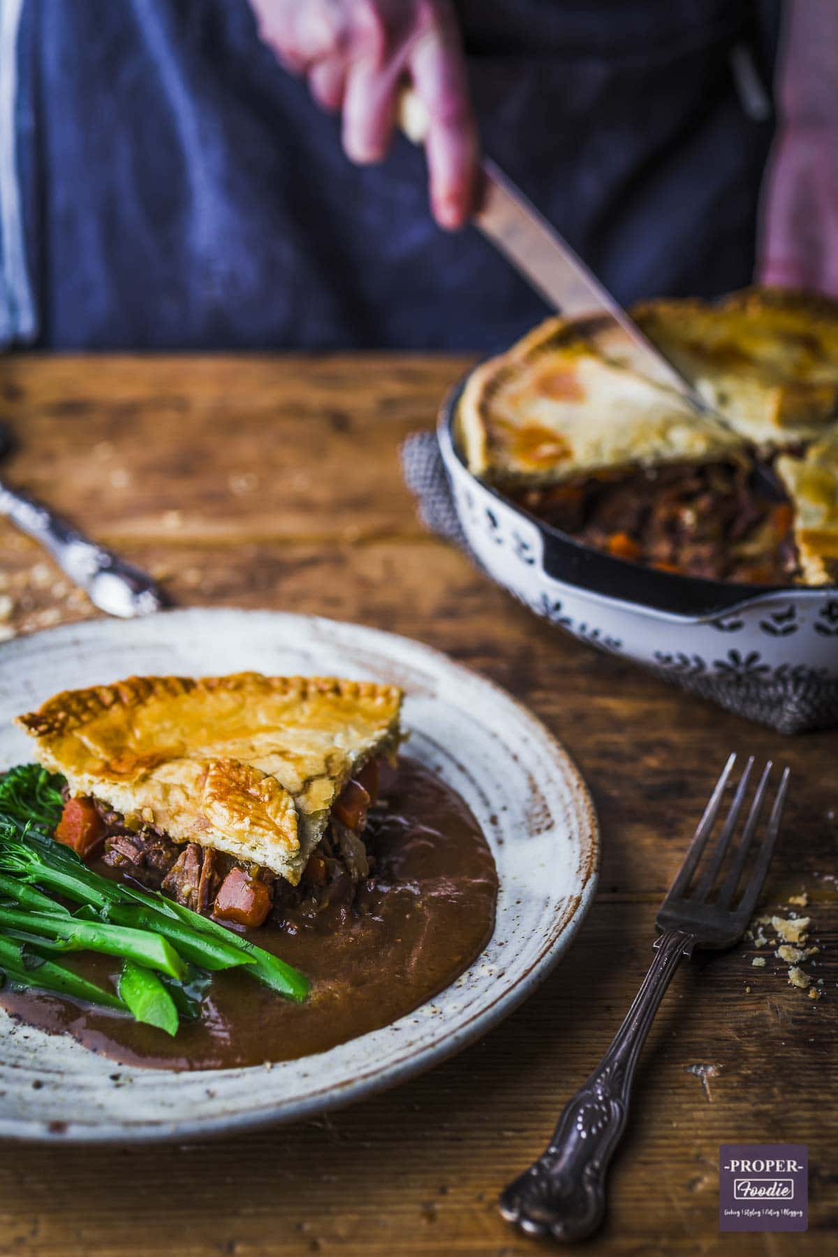 A slice of steak and ale pie on a plate with gravy and tender-stem broccoli with pie dish in the background.