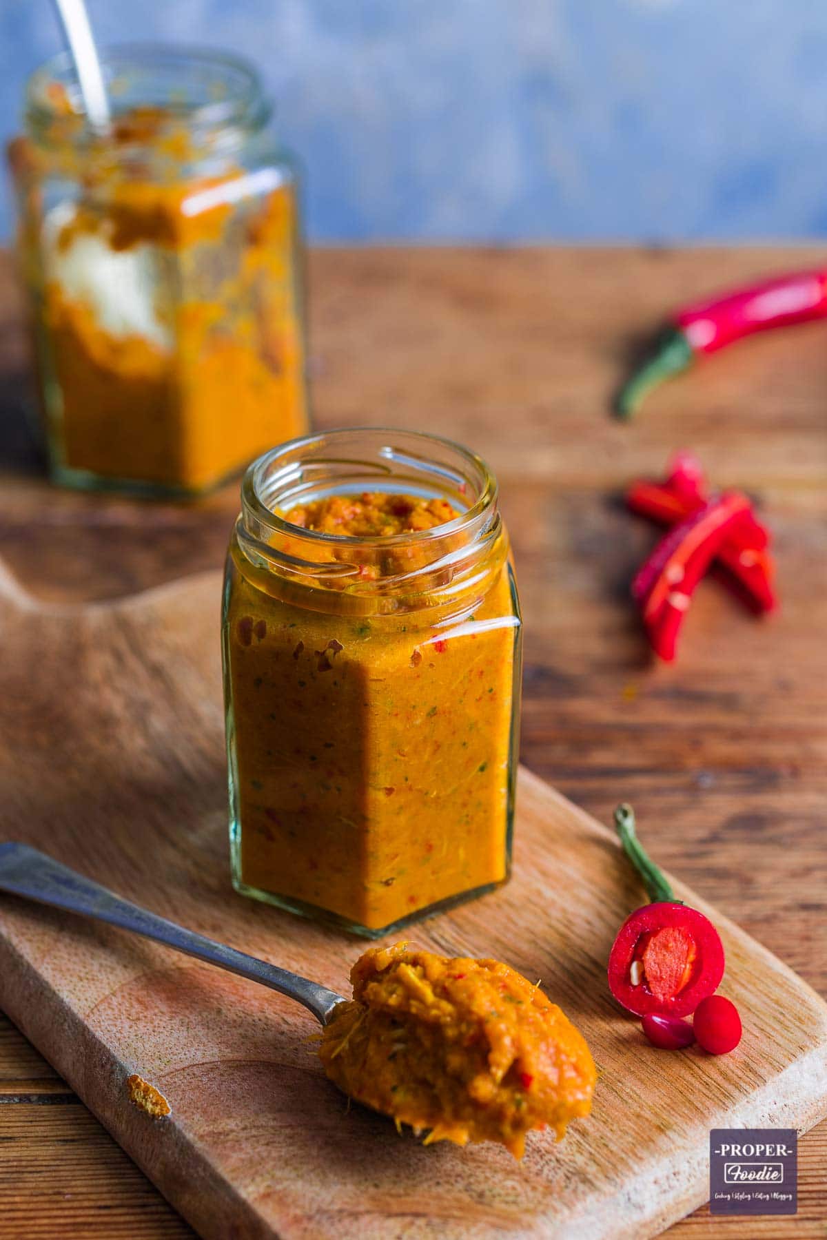 A small jar filled with homemade Thai red curry paste with a spoonful of paste in the foreground and a chopped up red chilli at the side.