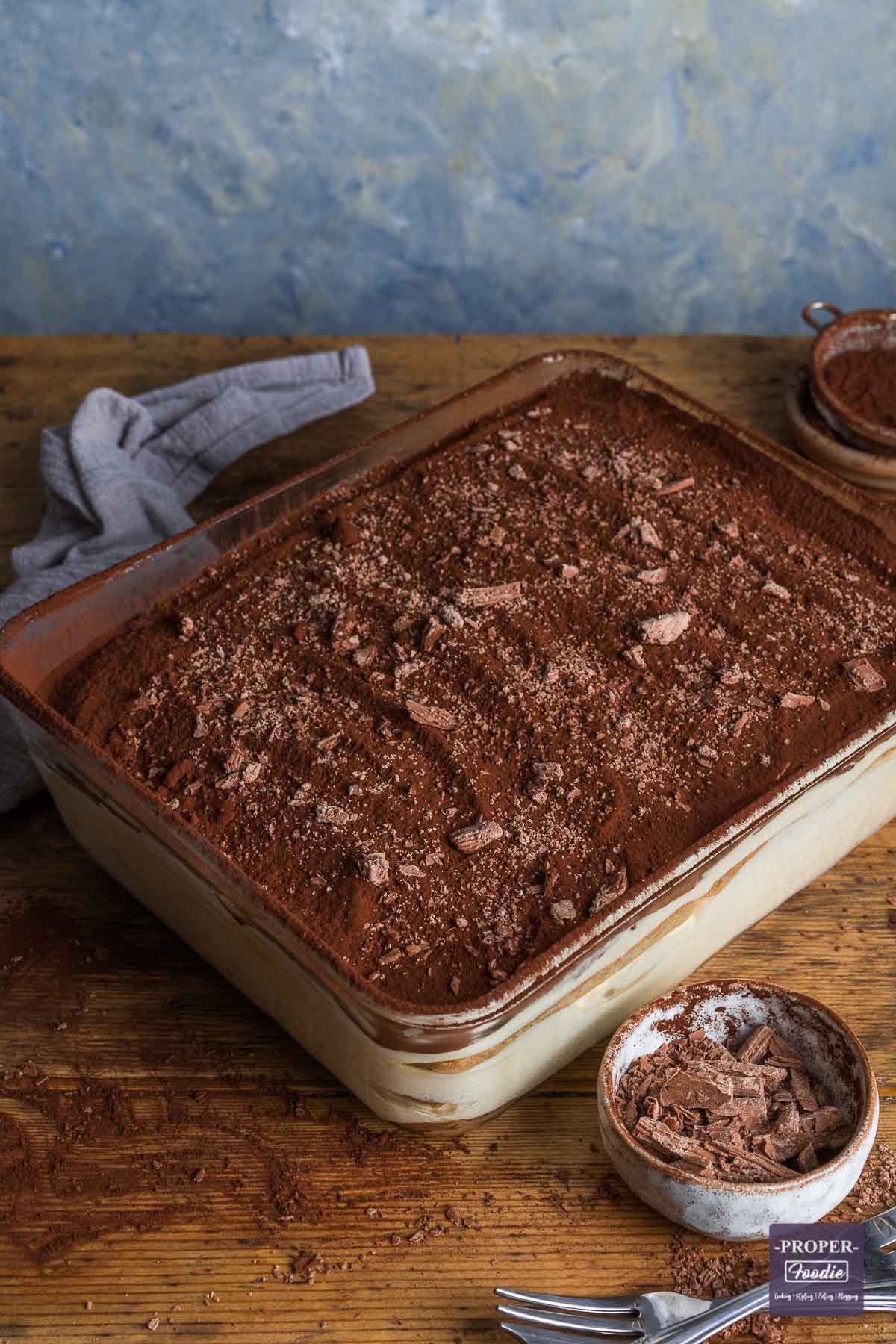 A large rectangular dish of traditional tiramisu topped with cocoa powder and flake chocolate with a small bowl of chocolate in the foreground