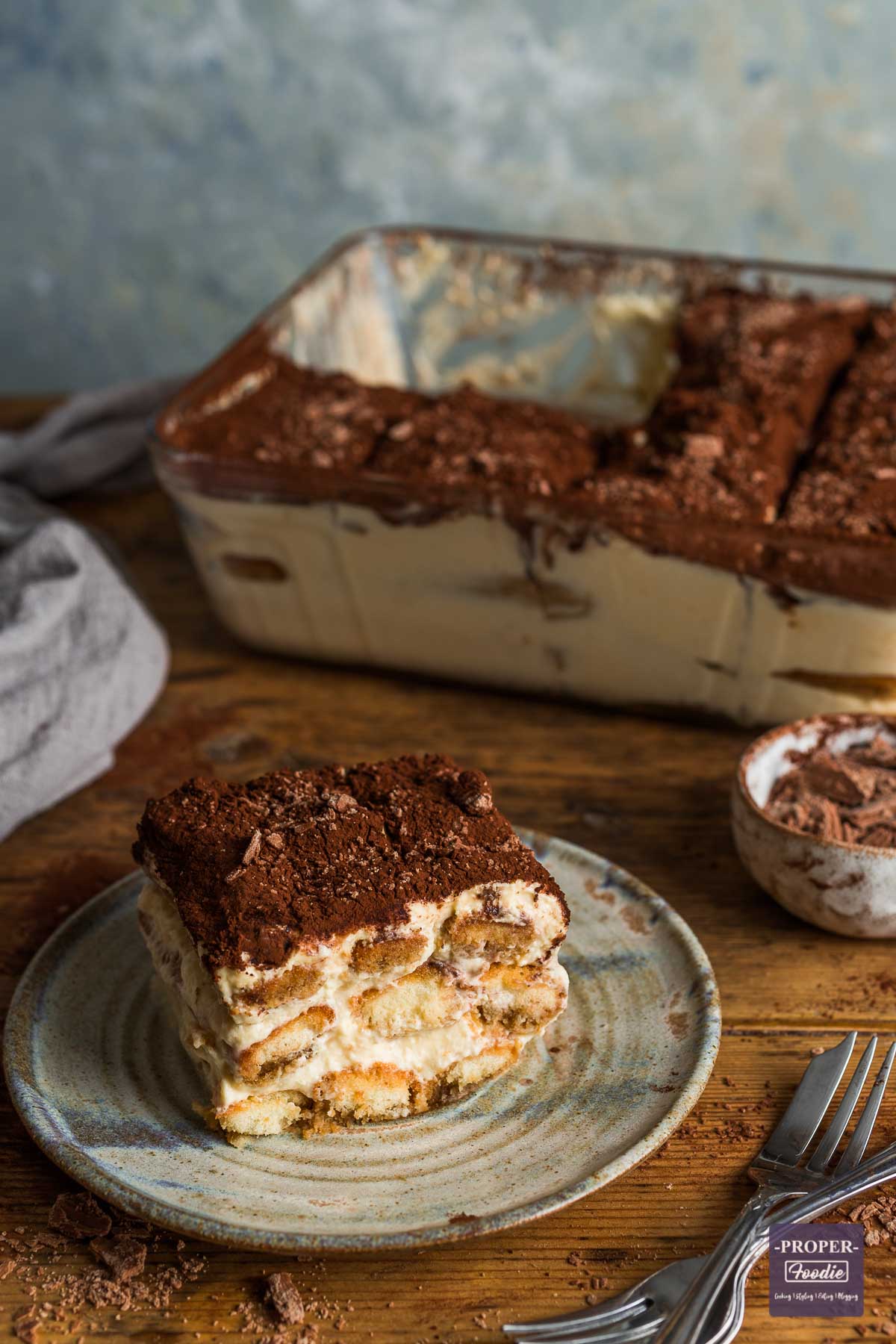 A small, square piece of tiramisu on a plate with the side view showing layers of sponge fingers and creamy mascarpone. The top is dusted with cocoa powder and a large dish of tiramisu is in the background.