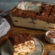 A small, square piece of tiramisu on a plate with the side view showing layers of sponge fingers and creamy mascarpone. The top is dusted with cocoa powder and a large dish of tiramisu is in the background.