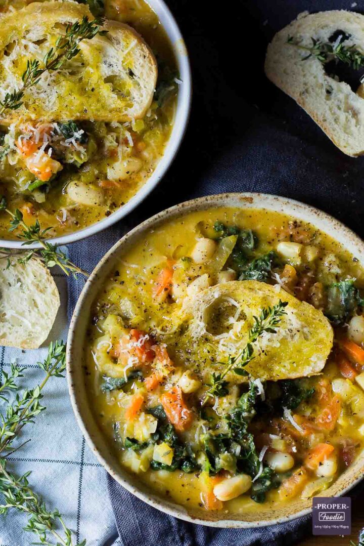 Ribollita vegetable and bean soup served up in two bowls and topped with crusty bread, grated parmesan and fresh sprigs of thyme.