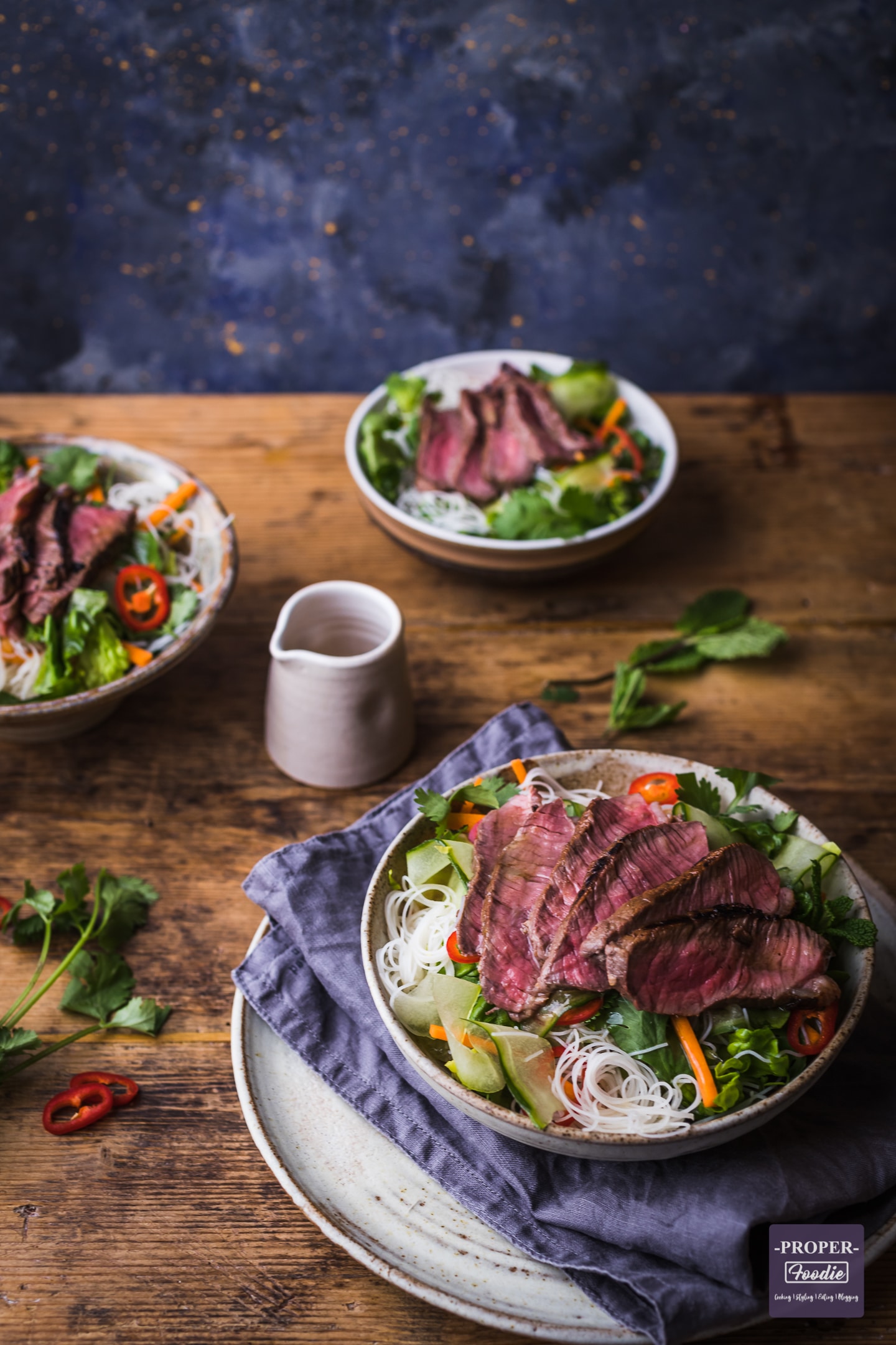 Beef noodle salad served up in 3 bowls with a small jug of salad dressing in the centre