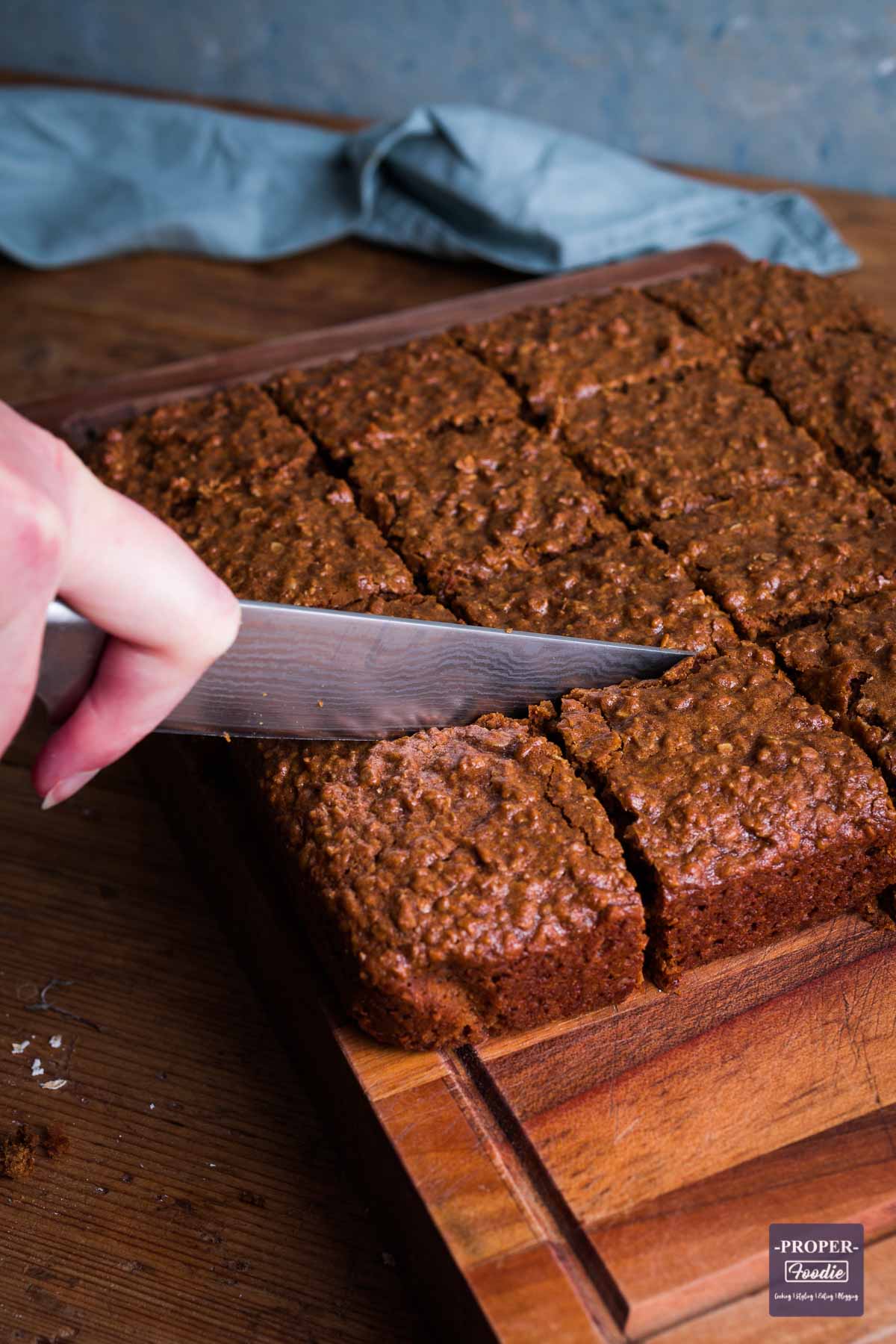 Full slab of parkin on a chopping board being sliced with a knife into 16 equal square pieces.